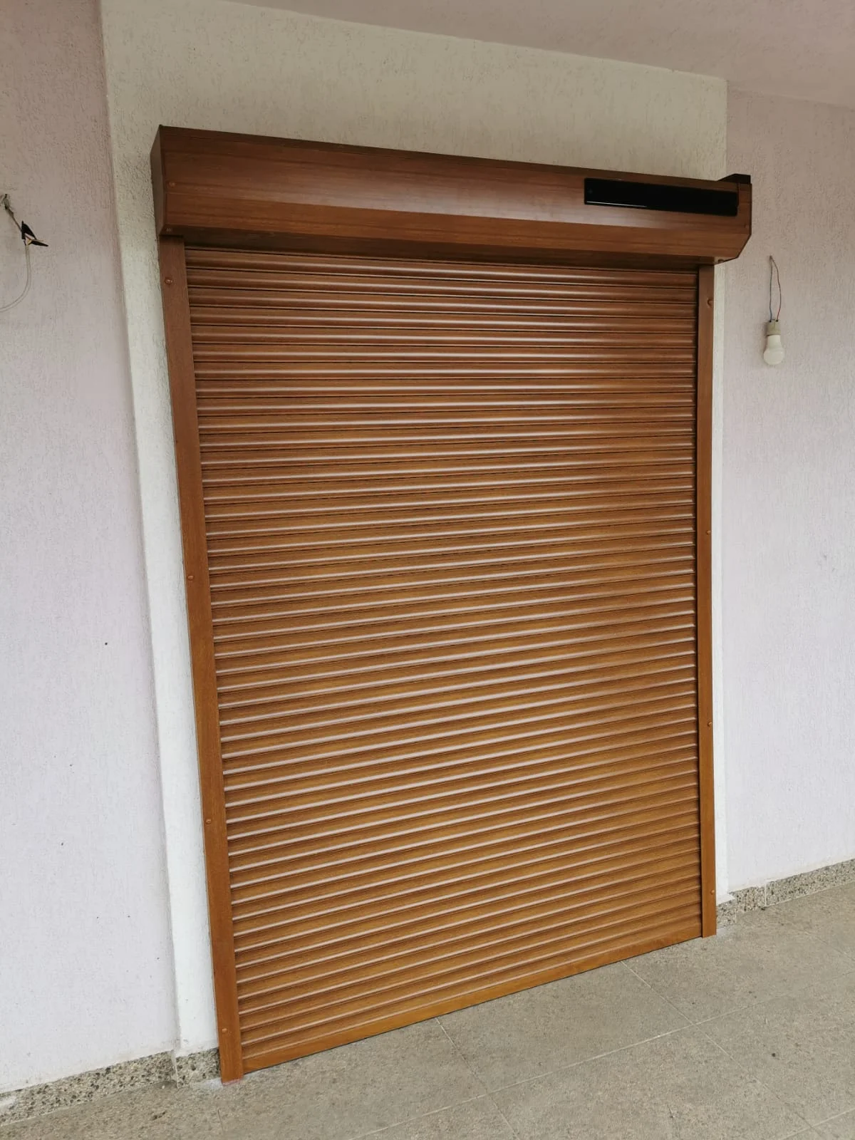 Rulou exterior din aluminiu în nuanță stejar auriu, montat deasupra unei uși, cu un mic panou solar pentru alimentarea motorului, în colțul casetei.